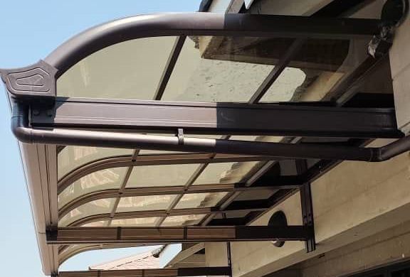A bronze-framed aluminium awning with a tinted roof panel, fitted at the corner of a house