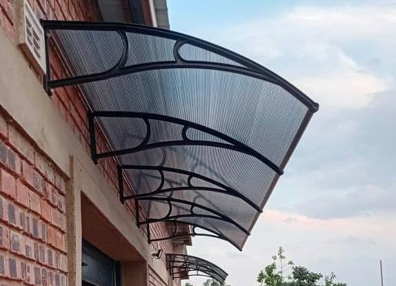 A large ribbed polycarbonate awning with graphite brackets mounted on a red brick wall against a blue sky