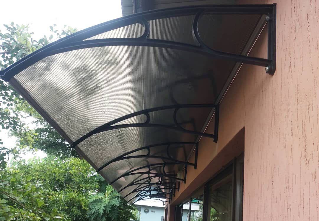 Underside view of a ribbed polycarbonate awning on curved graphite brackets above a glass door, with a garden below